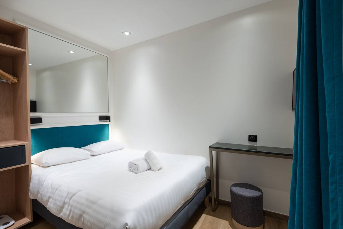 Una habitación de hotel con cama, escritorio, espejo y un taburete pequeño.