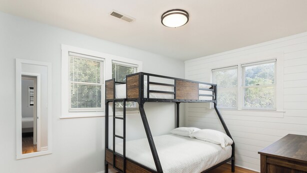 Una cama litera con estructura de madera y ropa de cama blanca en una habitación con paredes blancas y suelo de madera.