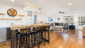 Una cocina amplia con una isla de madera y taburetes de bar, un área de comedor con un sofá gris y un ventanal con persianas.