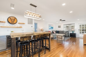 Una cocina amplia con una isla de madera y taburetes de bar, un área de comedor con un sofá gris y un ventanal con persianas.