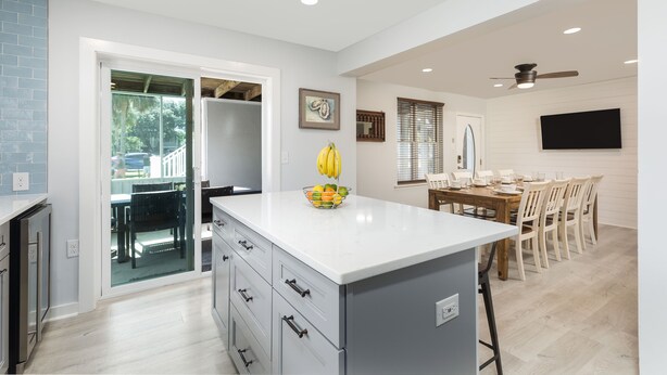 A modern kitchen with a central island, a dining area with a wooden table and chairs, and a view through a glass door to an outdoor patio.