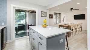 Una cocina moderna con una isla central, un comedor con mesa y sillas de madera, y un amplio ventanal con persianas.