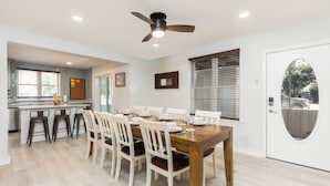 Un comedor con una mesa de madera dispuesta para seis personas, sillas blancas, un ventilador de techo y una cocina al fondo.