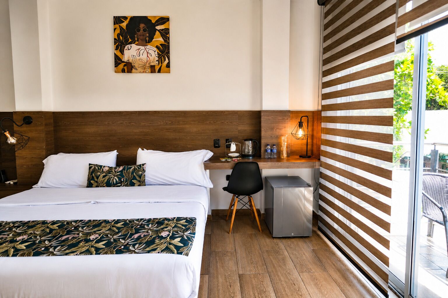Habitación de hotel con cama, escritorio, silla, basurero, lámpara de pared y ventana con persianas.