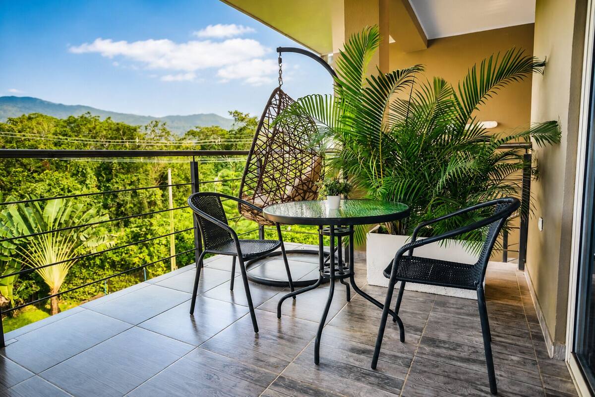 Een balkon met een ronde tafel en twee stoelen, omringd door bloempotten en uitzicht op groen.