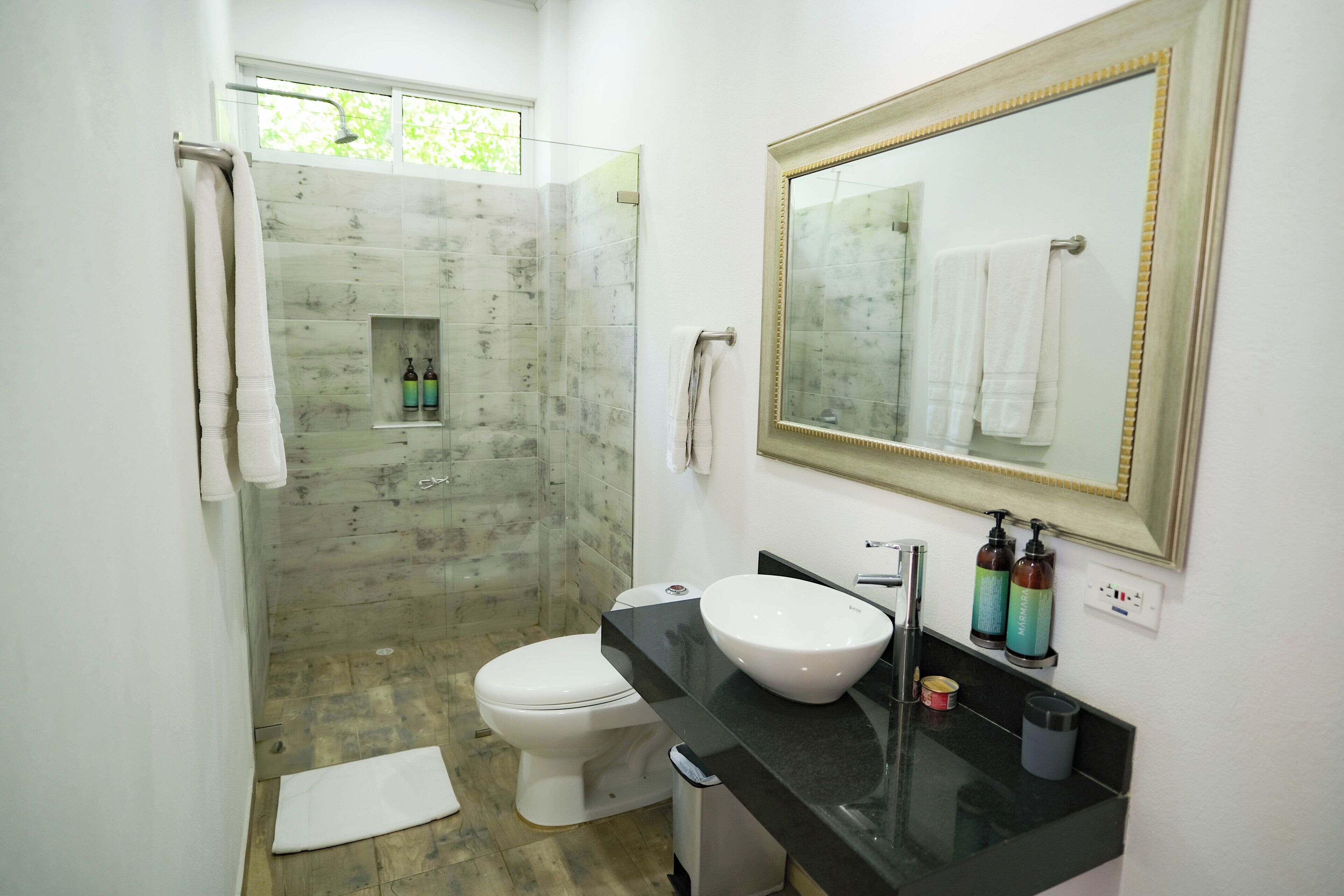 A bathroom with a glass-enclosed shower, a white toilet, a black countertop with a sink, and a mirror with a gold frame.