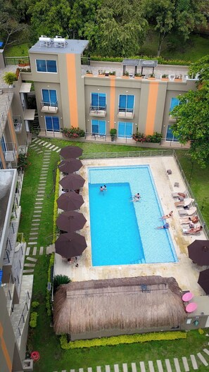 Ein Schwimmbecken mit Liegestühlen und Sonnenschirmen, umgeben von einem Gebäude mit Balkonen und einem Reetdach.