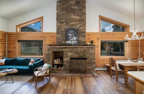 L’intérieur d’une cabane en rondins, avec une cheminée en pierre, des meubles en bois et de grandes fenêtres donnant sur le paysage extérieur.
