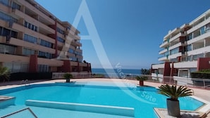 A swimming pool surrounded by multi-story buildings with balconies, a clear sky, and a view of the sea.