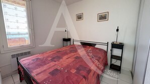 A bedroom with a bed, a bedside table, a nightstand, a window with blinds, and two framed pictures on the wall.