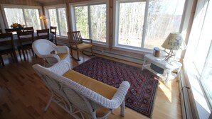 A cozy cabin interior with wooden floors, white wicker furniture, a red patterned rug, and large windows offering a view of trees.