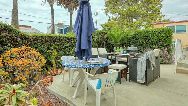 Eine Terrasse im Freien mit einem runden Tisch, Stühlen, einem blauen Sonnenschirm und einem Grill.