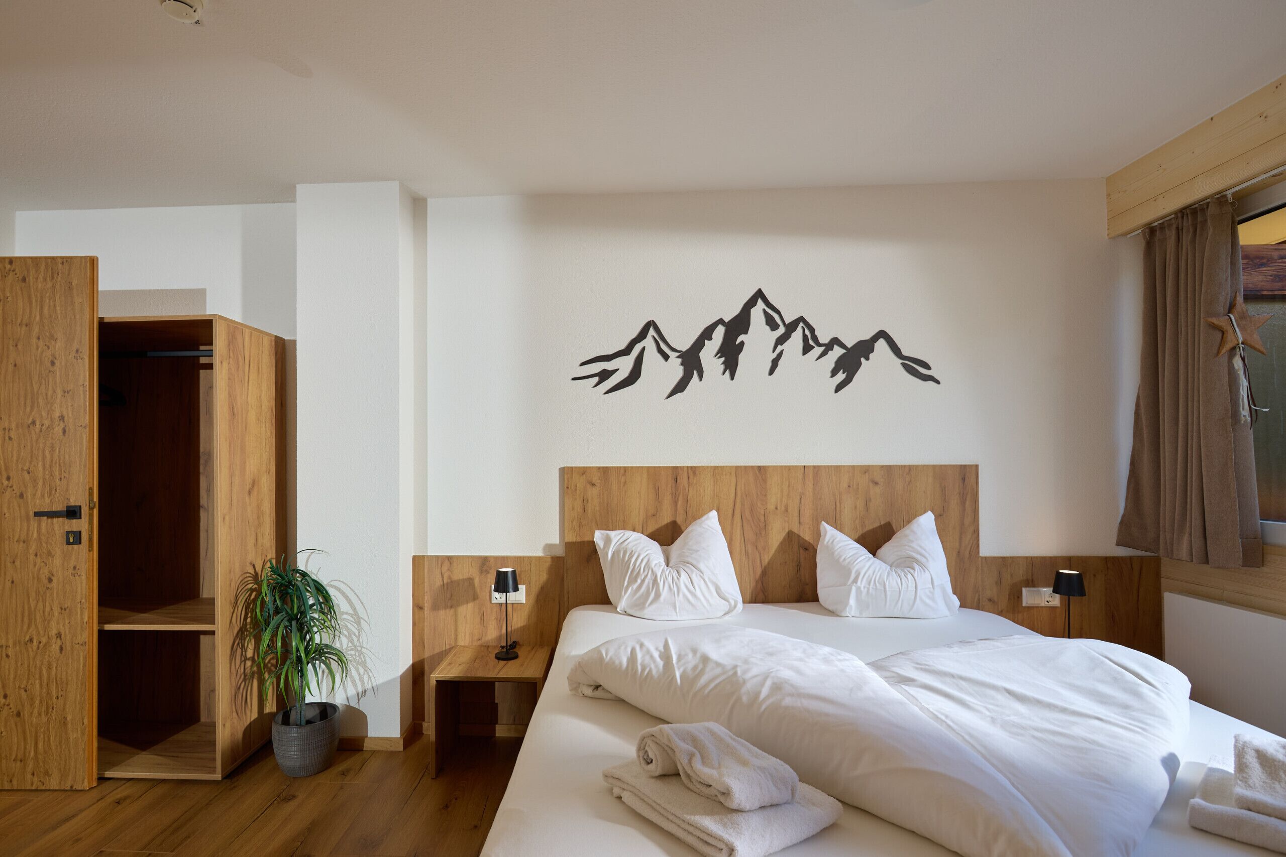 A hotel room with a wooden headboard, a bed with white linens, a nightstand with a lamp, a potted plant, and a wall-mounted mountain art piece.
