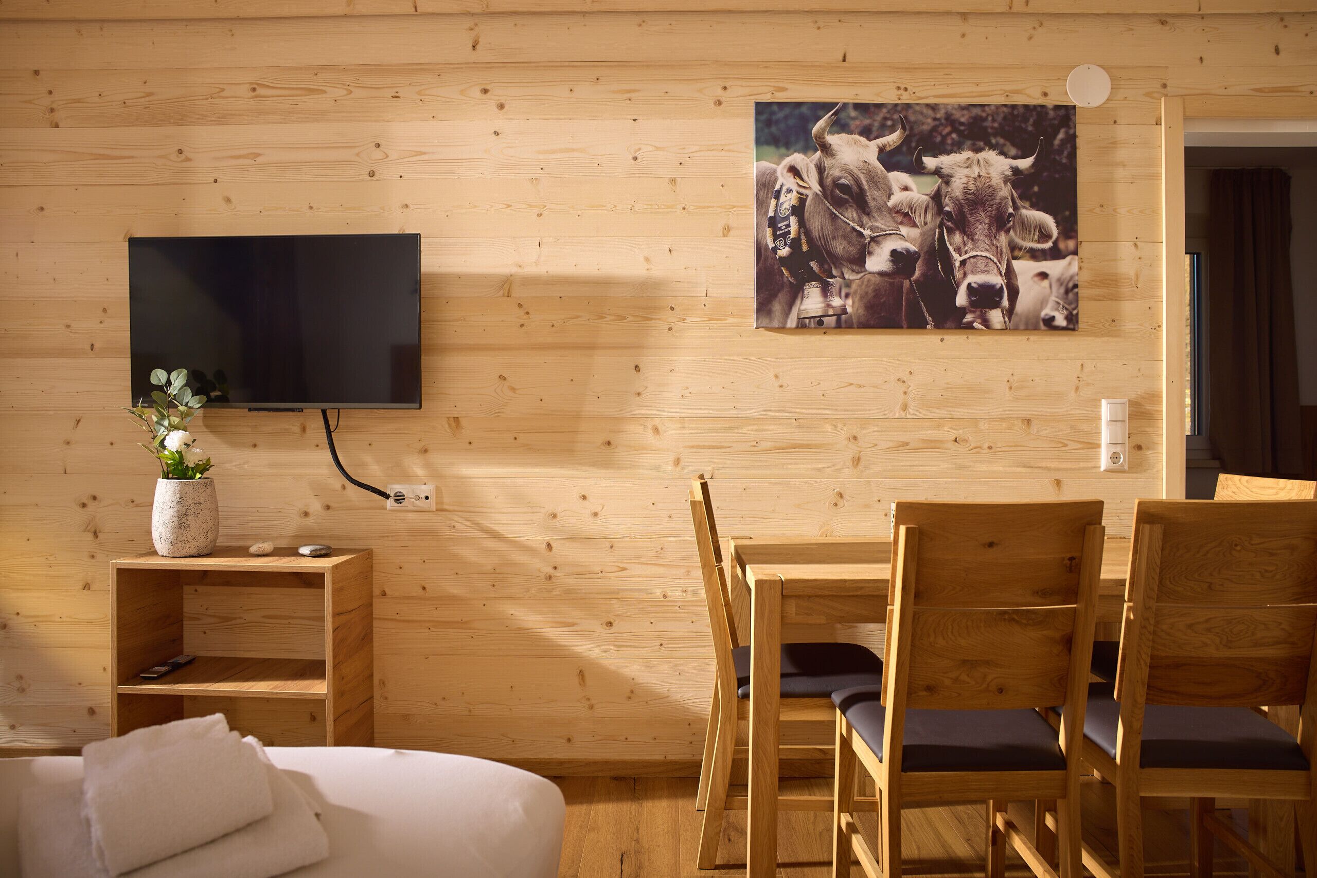 A wooden interior with a TV mounted on the wall, a dining table with chairs, and a bed with pillows.