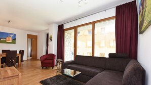 Een moderne woonkamer met een bruine bank, een glazen salontafel en een rode fauteuil. Er is een houten eettafel met stoelen en een klein bijzettafeltje. Grote ramen met rode gordijnen zorgen voor natuurlijk licht in de ruimte.