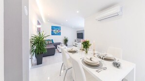 A modern dining area with a white table set for four, surrounded by white chairs. There is a potted plant on the left and a flat-screen TV mounted on the wall. - Villa Lioness in Kastel Luksic, playground & pool (Kastel Luksic)