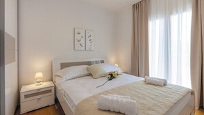 A neatly made bed with white linens and a bouquet of yellow flowers, a bedside table with a lamp, and a window with sheer curtains. - Villa Lioness in Kastel Luksic, playground & pool (Kastel Luksic)