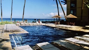 A pool area with lounge chairs and umbrellas, surrounded by palm trees and a view of the ocean. - Mahana 604 Oceanfront 1bd/1ba 1 Bedroom Condo by RedAwning (Lahaina)