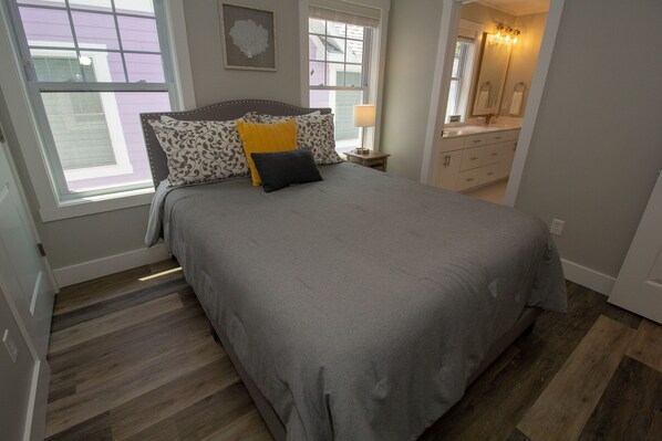 A bedroom with a large bed, a nightstand, a dresser, and a window with a view of a purple building.