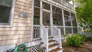 A two-story house with a porch, a visible number "112" sign, and a hose lying on the ground. - Charming, Spacious Resort Beach House for 12 (Michigan City)