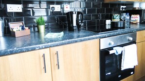 A modern kitchen with wooden cabinets, a black tiled backsplash, a coffee maker, and a bottle of Nutella on the counter. - Tudors eSuites Three Bedroom House (Coventry)