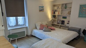 A bedroom with two beds, a window with a view, a bookshelf, and a desk.