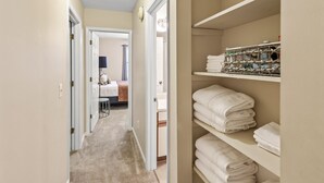 A hallway leading to a bedroom with a bed, a nightstand, and a lamp. There are shelves with folded towels and a metal rack with toiletries.