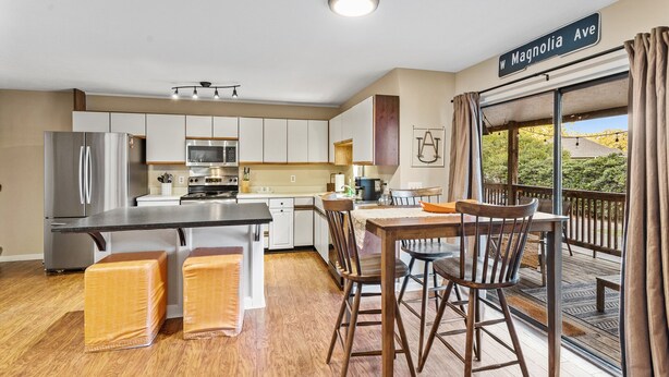 A modern kitchen with a stainless steel refrigerator, microwave, and stove, a dining area with wooden chairs and a table, and a view of a wooded area through a sliding glass door.