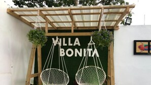 Una pérgola de madera con dos sillas de macramé colgando, una pared verde con el texto "VILLA BONITA" al fondo, y una obra de arte enmarcada a la derecha.