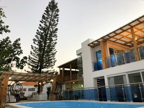 Un bâtiment moderne doté d’une piscine, d’une pergola en bois et de grands arbres.
