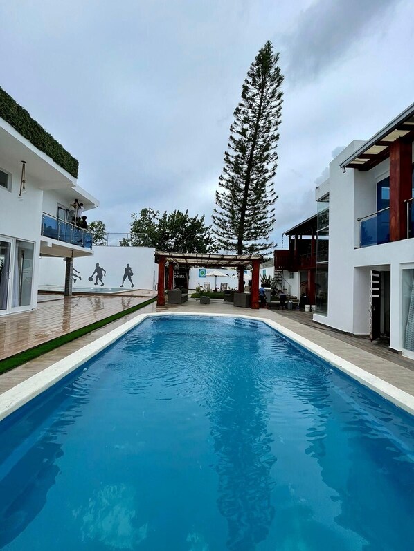 Un espace piscine moderne avec une piscine à l’eau d’un bleu limpide, entourée de bâtiments blancs dotés de balcons et d’un grand arbre au centre.