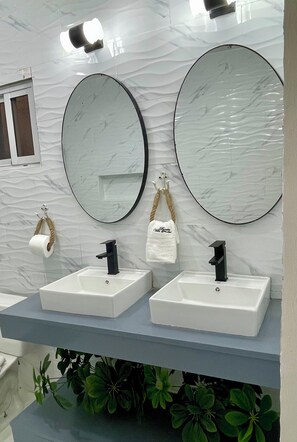 Une salle de bain avec deux lavabos, chacun doté d’un robinet noir, et deux grands miroirs ovales. Des serviettes sont suspendues à des crochets et un rouleau de papier toilette est fixé au mur.