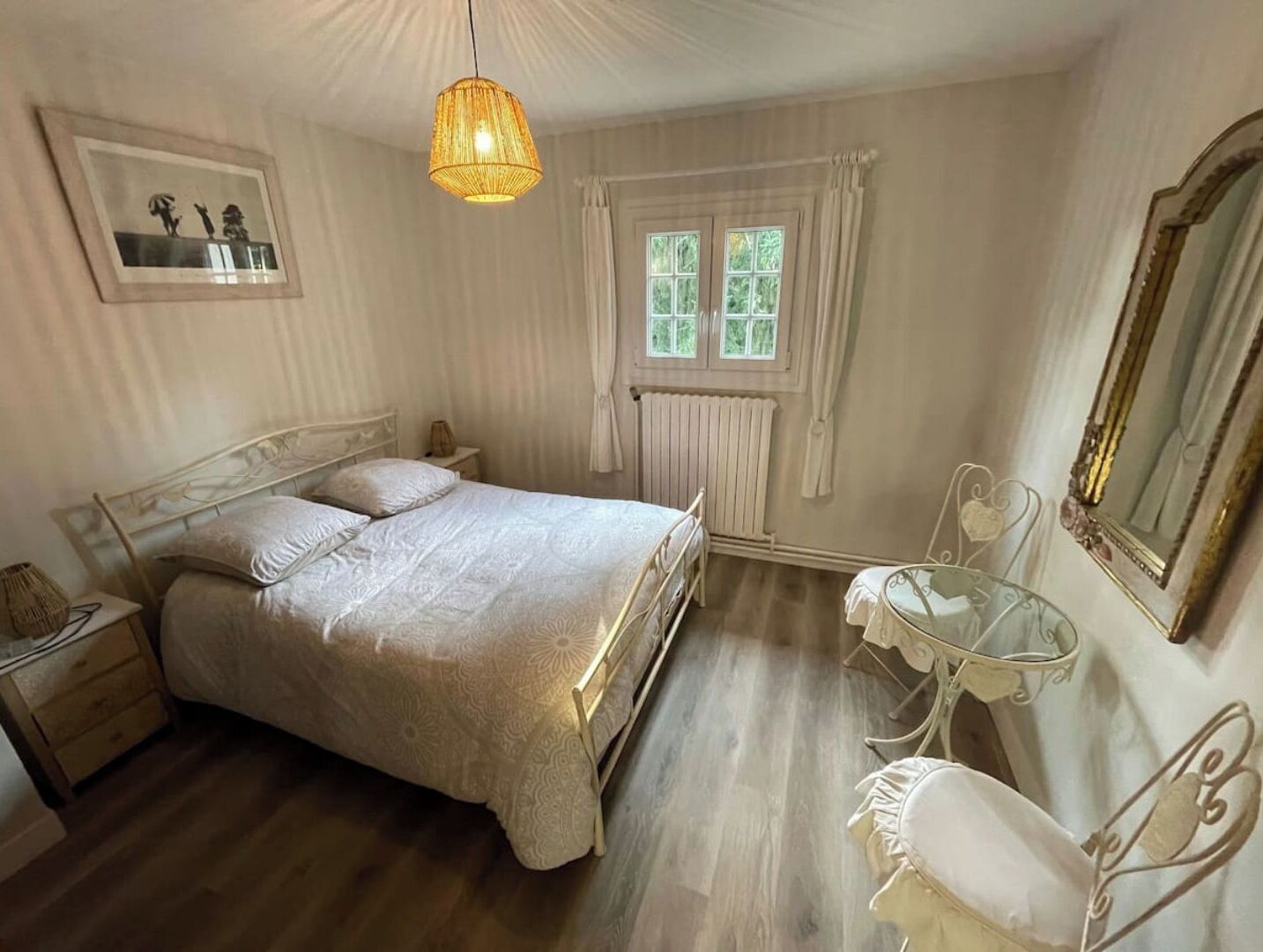 Une chambre à coucher comprenant un lit, des tables de chevet, une chaise, un luminaire suspendu, une fenêtre avec des rideaux et un tableau encadré accroché au mur.