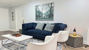 Una sala de estar con un sofá azul oscuro, sillones blancos, una mesa de centro blanca y un cuadro con una vista urbana enmarcada en la pared.