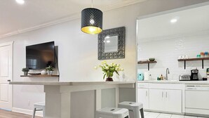 Una cocina moderna con armarios blancos, una isla central, taburetes de bar, una lámpara colgante, un televisor de pantalla plana y un espejo decorativo.