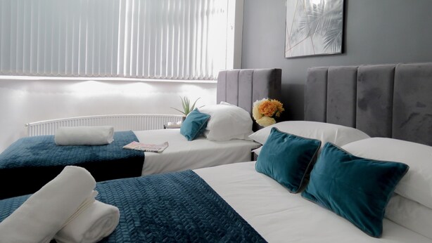 A modern hotel room with two beds, a grey headboard, a white radiator, and a window with blinds. - Tudors eSuites Four Bedroom House Garden (Coventry)
