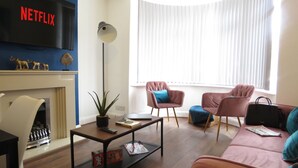 A modern living room with a fireplace, a Netflix sign, a floor lamp, and two armchairs. - Tudors eSuites Four Bedroom House Garden (Coventry)