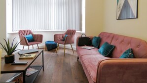 A living room with a pink sofa, two armchairs, a small table with a plant and books, and a window with white blinds. - Tudors eSuites Four Bedroom House Garden (Coventry)