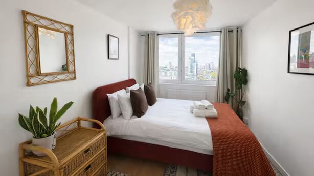 A bedroom with a bed, a hanging light fixture, a window with curtains, a plant, and a mirror on the wall. - Stylish flat near London Bridge with balcony views - Pass the Keys (London)
