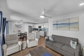 A modern living room with a grey sofa, armchairs, and a dining area with a kitchen in the background.