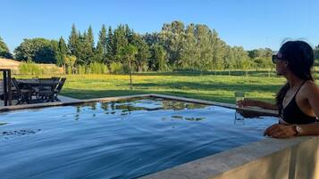 Une personne se détend au bord d’une piscine, avec vue sur un champ verdoyant et des arbres.