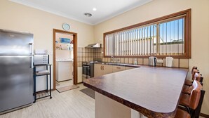 A modern kitchen with a large island, stainless steel appliances, and a window with vertical blinds. - Quota Cabin - Inlet Dreaming in a Fantastic Location (NAROOMA)