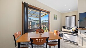 A dining area with a wooden table and chairs, a television, and a view of a house through a large window. - Quota Cabin - Inlet Dreaming in a Fantastic Location (NAROOMA)