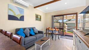 A living room with a grey sofa, blue cushions, a coffee table, and a TV. There's a dining area with a table and chairs, and a sliding glass door leading outside. - Quota Cabin - Inlet Dreaming in a Fantastic Location (NAROOMA)
