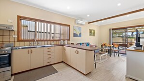 A modern kitchen with a built-in stove, a large window with blinds, and a dining area with a table and chairs. - Quota Cabin - Inlet Dreaming in a Fantastic Location (NAROOMA)