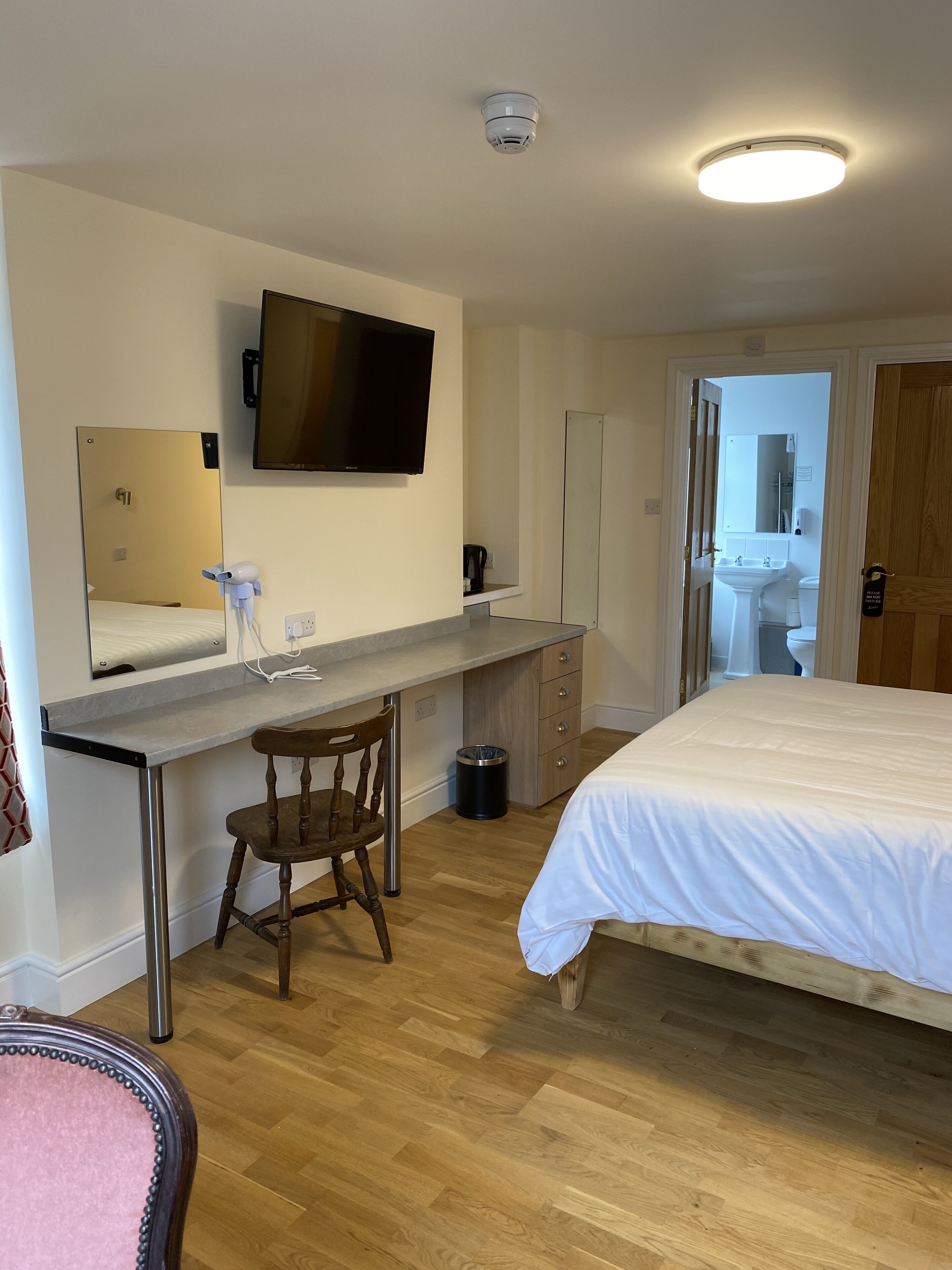 A hotel room with a bed, a desk with a chair, a TV mounted on the wall, a mirror, and a door leading to a bathroom.
