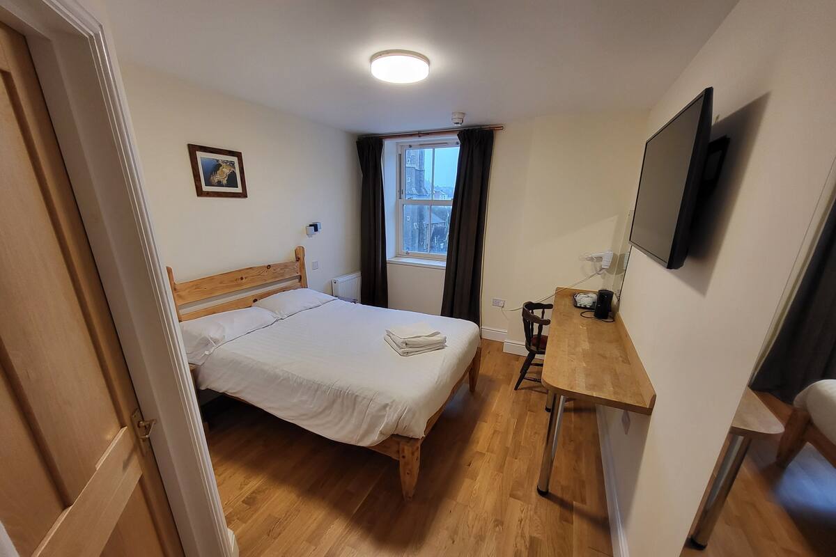 Ein Hotelzimmer mit einem hölzernen Bett, einem Schreibtisch, einem Sessel, einem Fernseher und einem Fenster mit Vorhängen.