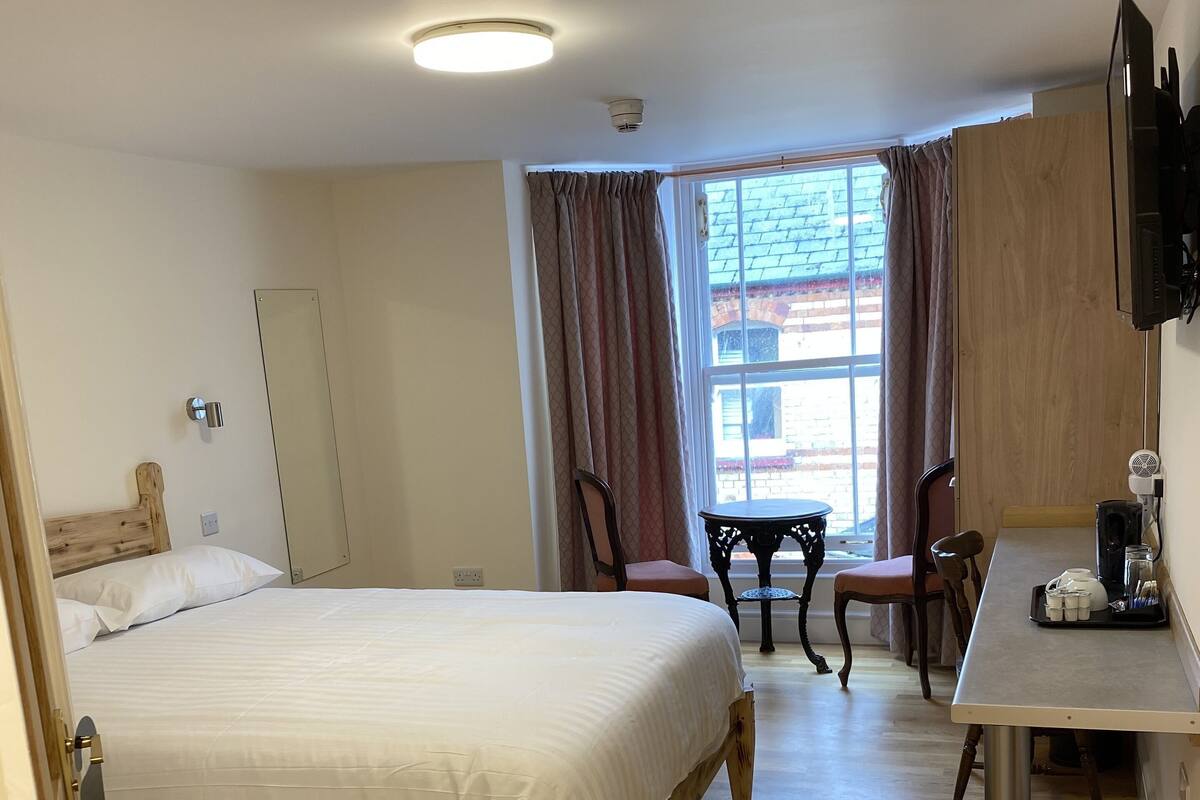 Ein Hotelzimmer mit einem hölzernen Bett, einem Schreibtisch mit Stuhl, einem Fenster mit Vorhängen und einem Fernseher.