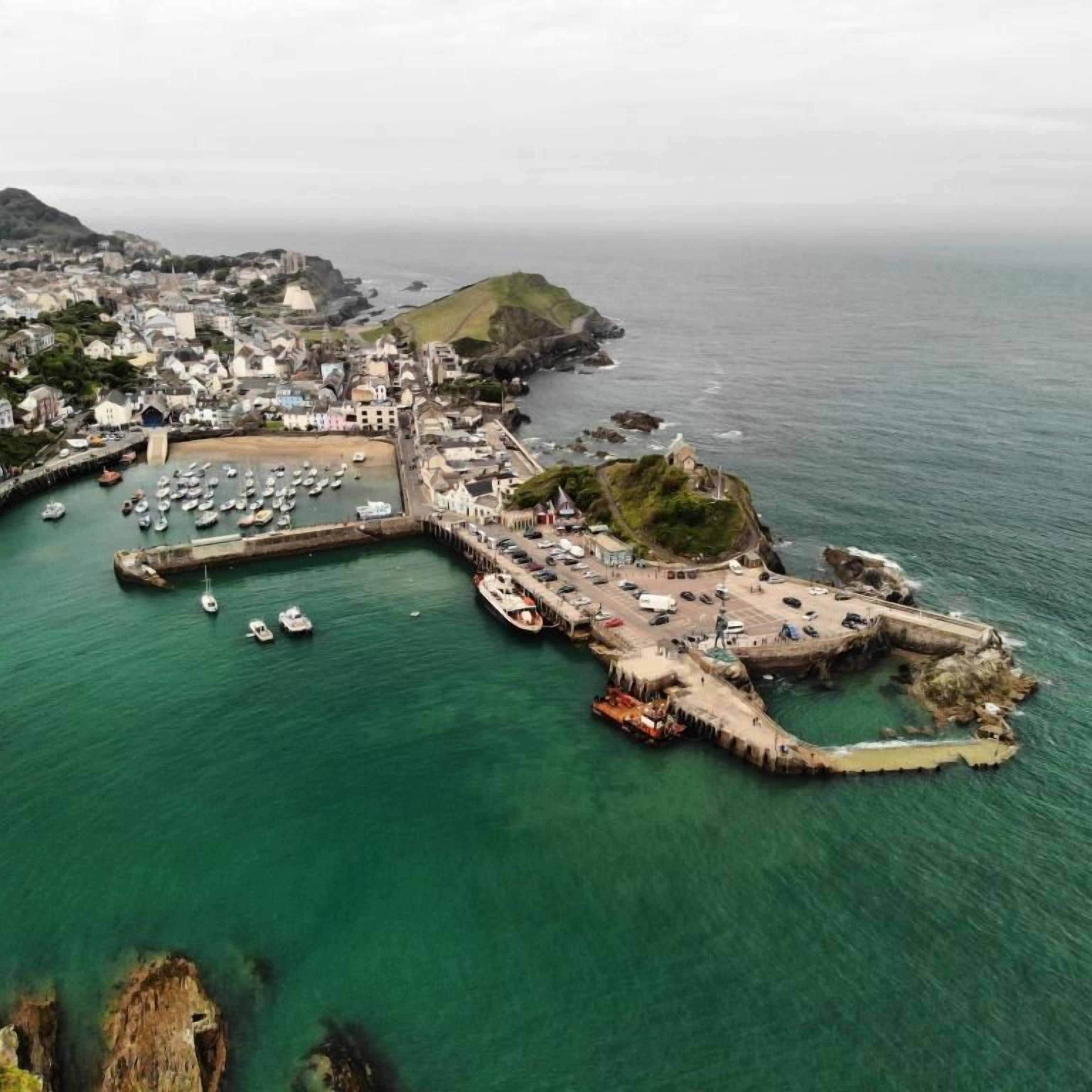 Vue aérienne d’une ville côtière avec un port de plaisance, des bâtiments et une colline imposante.