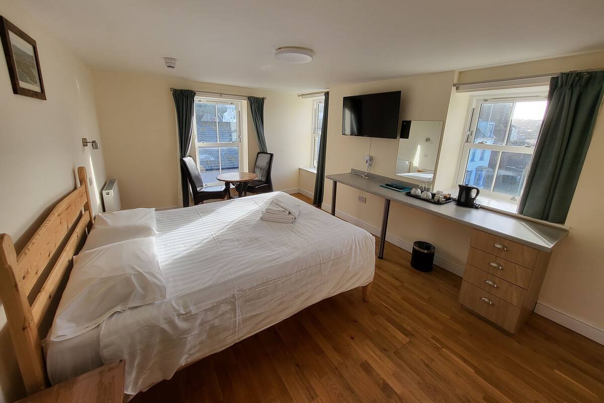 Ein Hotelzimmer mit einem Bett, einem Schreibtisch mit Flachbildfernseher, einem Sessel, einem kleinen Tisch und einem fenster mit Vorhängen.